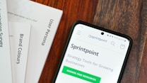 A wooden table with several documents and a smartphone. The documents are titled 'User Persona', 'Brand Colours', and 'Sprintpoint'. The smartphone displays a webpage for 'Sprintpoint', which offers strategy tools for growing businesses.