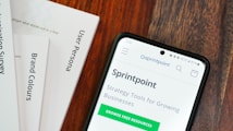 A wooden table with several documents and a smartphone. The documents are titled 'User Persona', 'Brand Colours', and 'Sprintpoint'. The smartphone displays a webpage for 'Sprintpoint', which offers strategy tools for growing businesses.