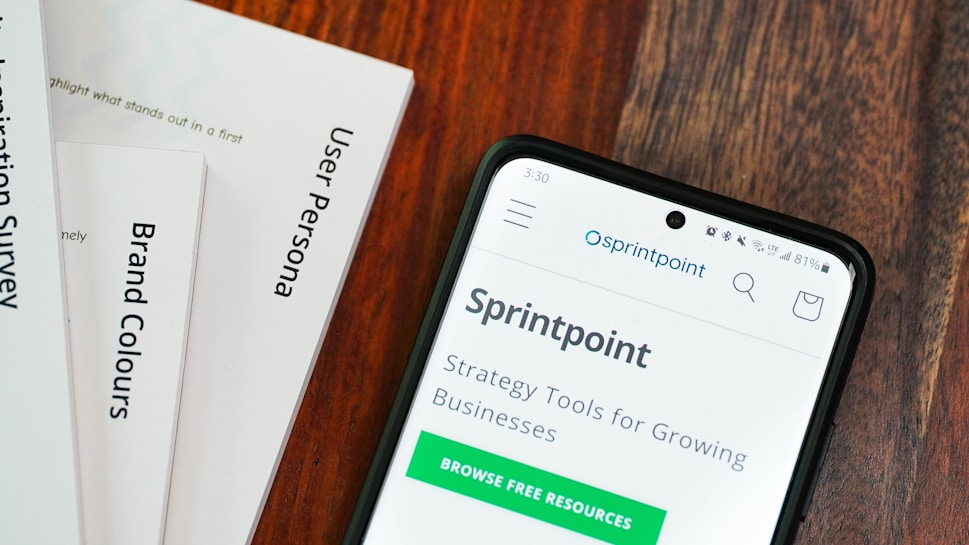 A wooden table with several documents and a smartphone. The documents are titled 'User Persona', 'Brand Colours', and 'Sprintpoint'. The smartphone displays a webpage for 'Sprintpoint', which offers strategy tools for growing businesses.