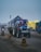 A tractor with a blue front and several lights is pulling a trailer covered with a white tarpaulin. There are colorful tents set up along the roadside. A motorbike is also visible behind the tractor, and traffic barriers line the road.