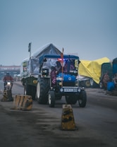 A tractor with a blue front and several lights is pulling a trailer covered with a white tarpaulin. There are colorful tents set up along the roadside. A motorbike is also visible behind the tractor, and traffic barriers line the road.