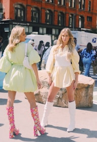 Two women stand outdoors, wearing short dresses with puffed sleeves. One wears a light green dress and the other a pale yellow dress. They both have long hair and are wearing knee-high boots. The background features a red brick building, a few vehicles, and several people walking by.