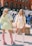 Two women stand outdoors, wearing short dresses with puffed sleeves. One wears a light green dress and the other a pale yellow dress. They both have long hair and are wearing knee-high boots. The background features a red brick building, a few vehicles, and several people walking by.