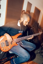 A person is sitting in a music studio playing an electric bass guitar. The individual has a beard and is wearing a dark sweater and jeans. The room is filled with guitar cases and acoustic panels on the walls, suggesting a professional music setting.