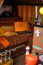 A cozy retro interior setup with a vintage orange record player placed on a plaid patterned seat. Surrounding elements include striped orange and yellow curtains, vibrant cushions, and a colorful knit throw. A small table features decorative items, and there is a crate of glass bottles on the floor.