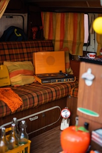 A cozy retro interior setup with a vintage orange record player placed on a plaid patterned seat. Surrounding elements include striped orange and yellow curtains, vibrant cushions, and a colorful knit throw. A small table features decorative items, and there is a crate of glass bottles on the floor.