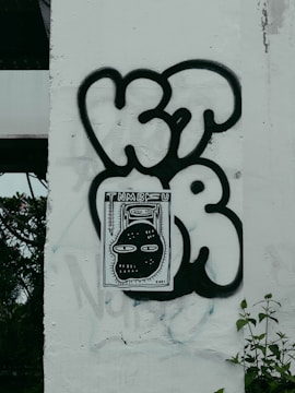 Graffiti art on a wall featuring bold black lettering and a central rectangular piece depicting a stylized cartoon face with large eyes. The wall has a light-colored background with some visible patches and foliage to the left.