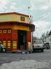 A small corner shop with vibrant red and yellow doors, located at the edge of a street. A motorbike is parked in front on one side, and an old van is parked on the other, hinting at a scene from an urban area. The shop has a traditional Chinese sign above the entrance. Overhead, numerous wires crisscross against a slightly cloudy sky.