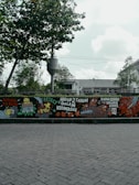 A mural painted on a wall features colorful and detailed illustrations, including animals like chickens and various objects. There are bold, handwritten phrases in a foreign language, adding a sense of urban art style. The mural is positioned along a street with a backdrop of trees and a cloudy sky.