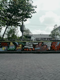 A mural painted on a wall features colorful and detailed illustrations, including animals like chickens and various objects. There are bold, handwritten phrases in a foreign language, adding a sense of urban art style. The mural is positioned along a street with a backdrop of trees and a cloudy sky.