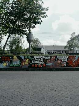 A mural painted on a wall features colorful and detailed illustrations, including animals like chickens and various objects. There are bold, handwritten phrases in a foreign language, adding a sense of urban art style. The mural is positioned along a street with a backdrop of trees and a cloudy sky.