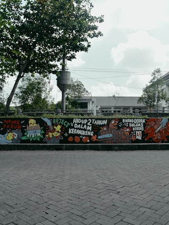 A mural painted on a wall features colorful and detailed illustrations, including animals like chickens and various objects. There are bold, handwritten phrases in a foreign language, adding a sense of urban art style. The mural is positioned along a street with a backdrop of trees and a cloudy sky.