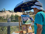 An artist wearing a blue shirt and white hat is painting a landscape on a wooden easel under a large blue umbrella. The scene includes sand dunes, green shrubs, and a clear blue sky. The painting captures the natural surroundings with vibrant colors and expressive brushstrokes.