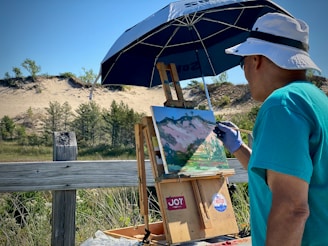 An artist wearing a blue shirt and white hat is painting a landscape on a wooden easel under a large blue umbrella. The scene includes sand dunes, green shrubs, and a clear blue sky. The painting captures the natural surroundings with vibrant colors and expressive brushstrokes.