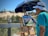 An artist wearing a blue shirt and white hat is painting a landscape on a wooden easel under a large blue umbrella. The scene includes sand dunes, green shrubs, and a clear blue sky. The painting captures the natural surroundings with vibrant colors and expressive brushstrokes.