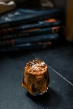 A glass filled with iced coffee sits on a dark surface, with frothy layers visible through the transparent sides. In the background, a stack of books with blurred titles adds a cozy, intellectual ambiance.