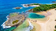Aerial view of a stunning beach with turquoise water, sandy shores, and rocky outcrops. Several horses and riders are visible on the sand. The surrounding area is lush with greenery, providing a stark contrast to the deep blue ocean.