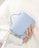 A hand is holding a light blue handbag against a white fabric background. The bag has a smooth texture and a simple design. A silver ring with a gemstone is worn on the hand, adding a touch of elegance.