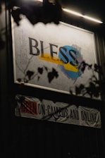 An illuminated sign with the word 'BLESS' in large, bold letters is displayed against a wall. The word is placed over abstract shapes in blue, yellow, and gray. Below the sign, a banner reads 'OPEN ON-CAMPUS AND ONLINE' with emphasis on the word 'OPEN' in red. The scene is partially obscured by dark silhouettes of leaves and branches.