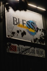 An illuminated sign with the word 'BLESS' in large, bold letters is displayed against a wall. The word is placed over abstract shapes in blue, yellow, and gray. Below the sign, a banner reads 'OPEN ON-CAMPUS AND ONLINE' with emphasis on the word 'OPEN' in red. The scene is partially obscured by dark silhouettes of leaves and branches.
