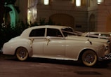 A classic white vintage car, adorned with ribbons, is parked on a city street at night. The car is illuminated by the soft glow of nearby streetlights and is positioned in front of a decorative building with balconies and lush greenery.