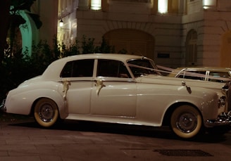 A classic white vintage car, adorned with ribbons, is parked on a city street at night. The car is illuminated by the soft glow of nearby streetlights and is positioned in front of a decorative building with balconies and lush greenery.