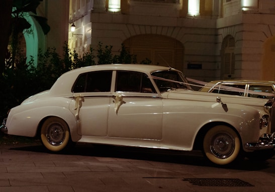 A classic white vintage car, adorned with ribbons, is parked on a city street at night. The car is illuminated by the soft glow of nearby streetlights and is positioned in front of a decorative building with balconies and lush greenery.