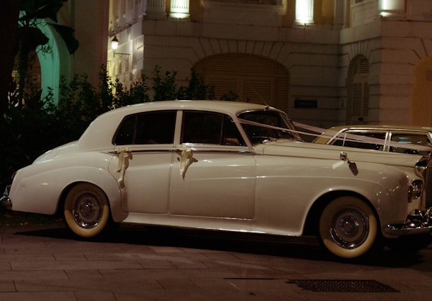 A classic white vintage car, adorned with ribbons, is parked on a city street at night. The car is illuminated by the soft glow of nearby streetlights and is positioned in front of a decorative building with balconies and lush greenery.