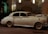 A classic white vintage car, adorned with ribbons, is parked on a city street at night. The car is illuminated by the soft glow of nearby streetlights and is positioned in front of a decorative building with balconies and lush greenery.