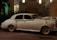 A classic white vintage car, adorned with ribbons, is parked on a city street at night. The car is illuminated by the soft glow of nearby streetlights and is positioned in front of a decorative building with balconies and lush greenery.