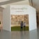 A small exhibition booth featuring numerous photos displayed on the walls. The booth has a white exterior with Thai and English text reading 'Life@Home.' Two individuals stand in front of the booth, looking inside.