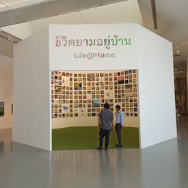 A small exhibition booth featuring numerous photos displayed on the walls. The booth has a white exterior with Thai and English text reading 'Life@Home.' Two individuals stand in front of the booth, looking inside.