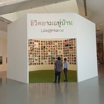 A small exhibition booth featuring numerous photos displayed on the walls. The booth has a white exterior with Thai and English text reading 'Life@Home.' Two individuals stand in front of the booth, looking inside.