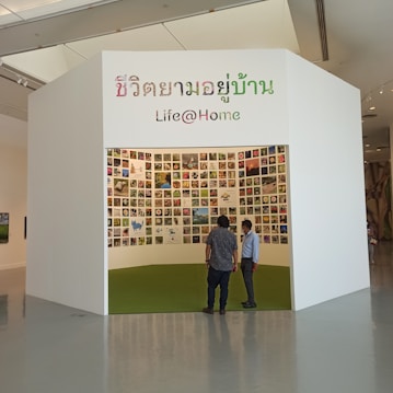 A small exhibition booth featuring numerous photos displayed on the walls. The booth has a white exterior with Thai and English text reading 'Life@Home.' Two individuals stand in front of the booth, looking inside.