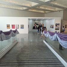 An art gallery with a sleek modern interior featuring several framed artworks displayed on white walls. The gallery is adorned with purple and white drapery along the glass railings. Several people are walking through the space, casually observing the artwork.