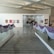 An art gallery with a sleek modern interior featuring several framed artworks displayed on white walls. The gallery is adorned with purple and white drapery along the glass railings. Several people are walking through the space, casually observing the artwork.