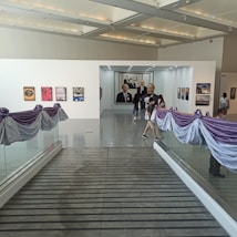 An art gallery with a sleek modern interior featuring several framed artworks displayed on white walls. The gallery is adorned with purple and white drapery along the glass railings. Several people are walking through the space, casually observing the artwork.