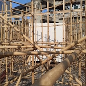 A complex structure of bamboo scaffolding set against a partially constructed concrete building. The sunlight casts intricate shadows on the bamboo, highlighting the geometric patterns created by the intersecting poles. In the background, multiple floors of the unfinished building with large windows are visible.