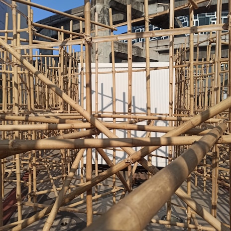 A complex structure of bamboo scaffolding set against a partially constructed concrete building. The sunlight casts intricate shadows on the bamboo, highlighting the geometric patterns created by the intersecting poles. In the background, multiple floors of the unfinished building with large windows are visible.