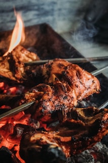 Pieces of grilled meat with a charred texture are being cooked over red-hot coals. Flames lick at the surface, and metal skewers pierce through the thick meat.