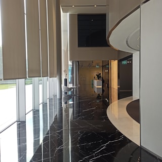 A modern interior space featuring large vertical window blinds on the left with natural light streaming in. The black marble floor reflects the surroundings, adding an elegant touch. On the right, there is a curved wall with a light strip feature. In the background, a reception area with a sleek white desk and some decorative plants is visible.