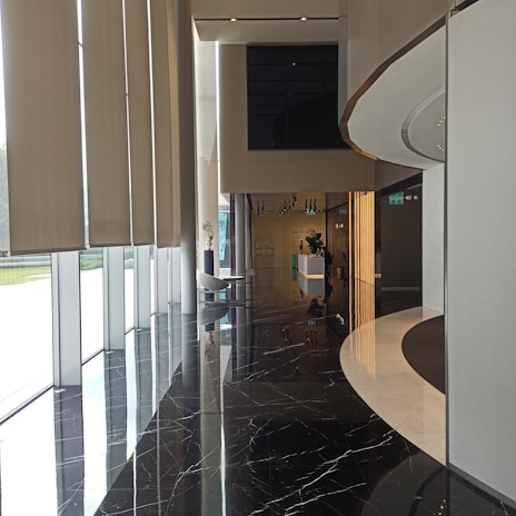 A modern interior space featuring large vertical window blinds on the left with natural light streaming in. The black marble floor reflects the surroundings, adding an elegant touch. On the right, there is a curved wall with a light strip feature. In the background, a reception area with a sleek white desk and some decorative plants is visible.
