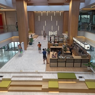 A modern indoor area with wooden elements and marble flooring. There are multiple levels of seating with green cushions and steps leading up to a cafe counter. A person stands near the counter while others walk around or take photographs. Potted plants and a colorful abstract painting on an easel add to the decor. A large sign with the word 'Brave' is illuminated on the wall behind a small stage area.