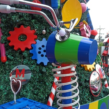 A colorful arrangement with large, decorative gears and springs against a background of artificial greenery. Brightly colored mechanical parts, like a blue and green cylindrical object with attached handlebars, are featured prominently amidst red, yellow, and blue gears. There are also reflective surfaces and candy cane-like striped elements.