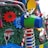 A colorful arrangement with large, decorative gears and springs against a background of artificial greenery. Brightly colored mechanical parts, like a blue and green cylindrical object with attached handlebars, are featured prominently amidst red, yellow, and blue gears. There are also reflective surfaces and candy cane-like striped elements.