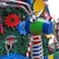 A colorful arrangement with large, decorative gears and springs against a background of artificial greenery. Brightly colored mechanical parts, like a blue and green cylindrical object with attached handlebars, are featured prominently amidst red, yellow, and blue gears. There are also reflective surfaces and candy cane-like striped elements.