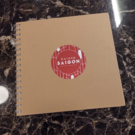 A spiral-bound notebook with a plain brown cover is placed on a dark marble surface. The cover features a red circular sticker in the center with the text 'Maison Saigon' and a decorative white geometric pattern.