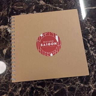 A spiral-bound notebook with a plain brown cover is placed on a dark marble surface. The cover features a red circular sticker in the center with the text 'Maison Saigon' and a decorative white geometric pattern.