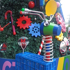 A creative and colorful arrangement featuring painted gears and a whimsical abstract sculpture resembling a bicycle. The setup incorporates bright elements like red and blue gears, a green and blue cylinder with handlebars and mirrors, and a spring-loaded red and silver base. The background consists of dark green faux foliage, adding contrast to the vibrant colors.