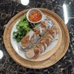 A woven bamboo tray contains a plate of Asian cuisine with fresh vegetables, steamed rolls topped with crispy fried onions, and sliced sausages arranged neatly. A small bowl of dipping sauce with red chilies sits next to the main dish.